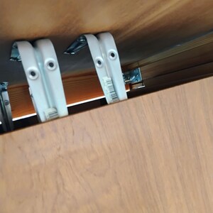 Op de afbeelding: Twee witte metalen L-vormige beugels zijn gemonteerd aan de onderkant van een lichtbruin houten oppervlak. De beugels zijn voorzien van ronde schroefgaten en een kleine barcode-sticker aan de zijkant, waarmee het houten paneel is bevestigd.