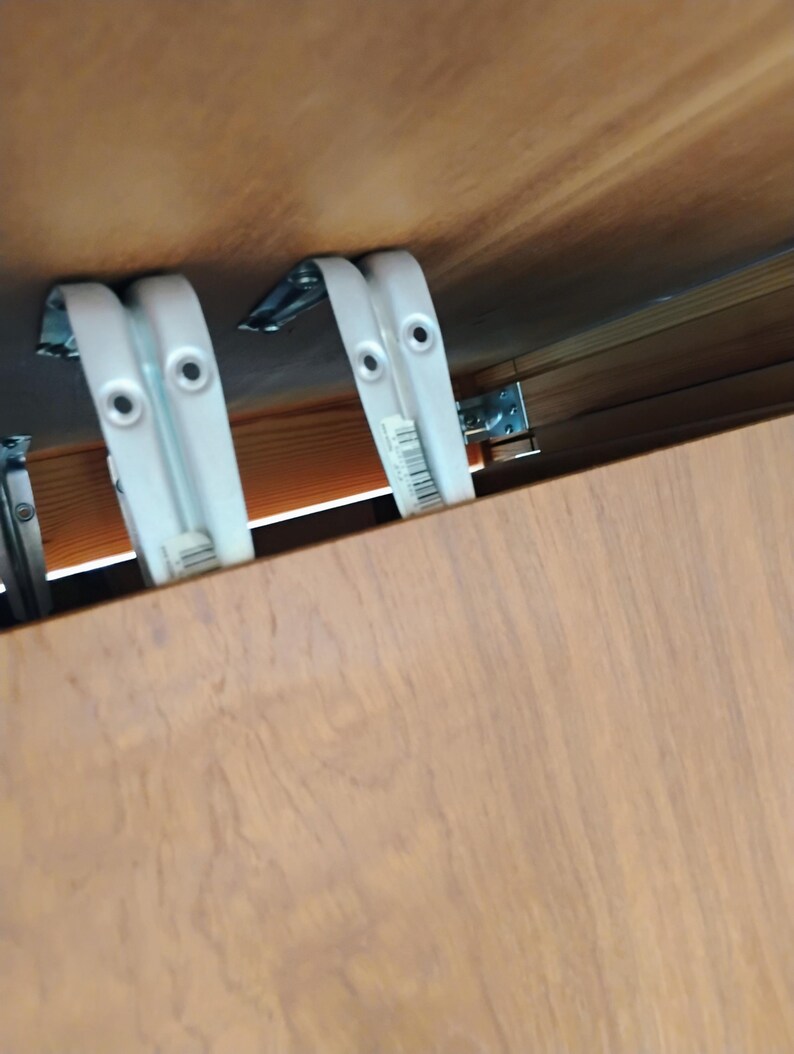 Op de afbeelding: Twee witte metalen L-vormige beugels zijn gemonteerd aan de onderkant van een lichtbruin houten oppervlak. De beugels zijn voorzien van ronde schroefgaten en een kleine barcode-sticker aan de zijkant, waarmee het houten paneel is bevestigd.