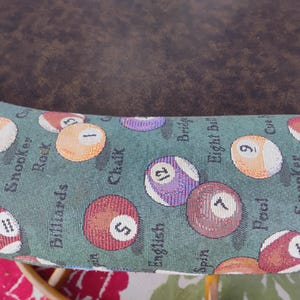 May include: Close-up of a section of a pool table. The fabric is green with images of billiard balls and words like "Pool", "Snooker", and "Billiards". The balls are numbered and colored.