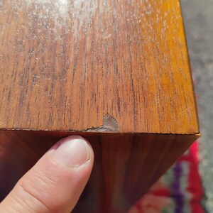 Op de afbeelding: Een rechthoekig houten object met een zichtbaar houtnerfpatroon in een medium bruine tint. Het oppervlak vertoont een klein gebied met afbladderende afwerking of schade langs de rand, waardoor een donkerbruine laag onder het fineer zichtbaar is.