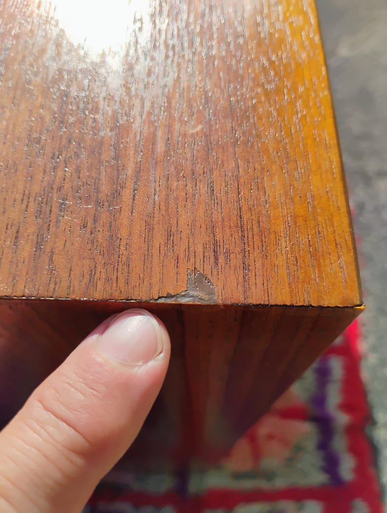 Op de afbeelding: Een rechthoekig houten object met een zichtbaar houtnerfpatroon in een medium bruine tint. Het oppervlak vertoont een klein gebied met afbladderende afwerking of schade langs de rand, waardoor een donkerbruine laag onder het fineer zichtbaar is.