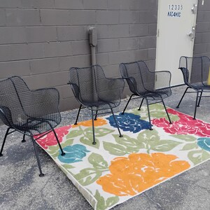 May include: Four black metal chairs with a mesh design are arranged on a white rug with a floral pattern. The rug has red, orange, blue, and green flowers.