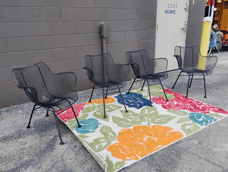 May include: Four black metal chairs with a mesh design are arranged on a white rug with a floral pattern. The rug has red, orange, blue, and green flowers.
