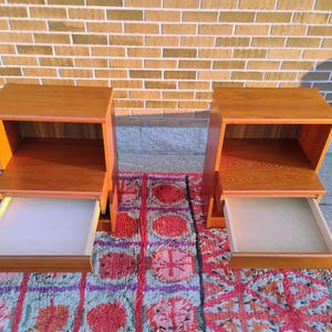 May include: Two wooden nightstands with drawers. The nightstands are made of light brown wood and have a simple, modern design. The drawers are open, revealing a white interior. The nightstands are placed on a colorful rug.