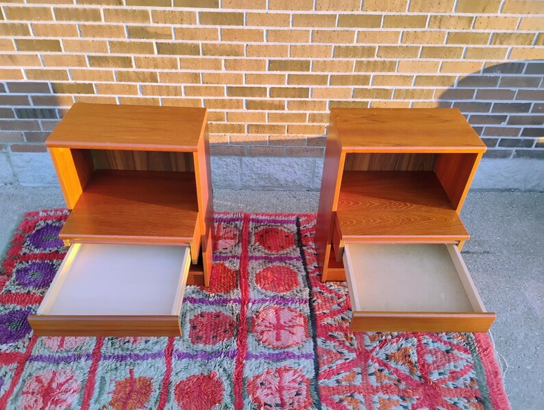 May include: Two wooden nightstands with drawers. The nightstands are made of light brown wood and have a simple, modern design. The drawers are open, revealing a white interior. The nightstands are placed on a colorful rug.