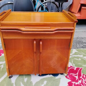 May include: A wooden bar cart with a drop-down serving tray, two doors, and wheels. The cart is made of light brown wood with a natural finish.