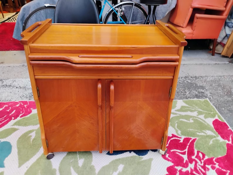May include: A wooden bar cart with a drop-down serving tray, two doors, and wheels. The cart is made of light brown wood with a natural finish.