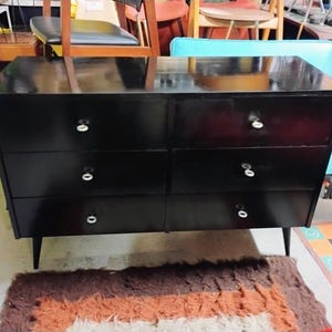 May include: A black dresser with six drawers and white knobs. The dresser has a glossy finish and sits on four tapered legs. The furniture piece is a mid-century modern style.