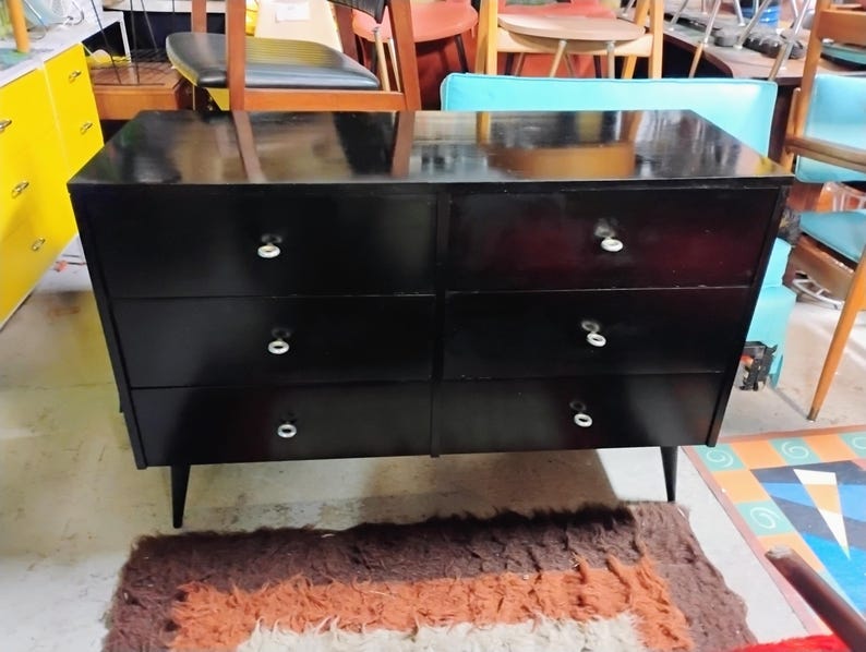 May include: A black dresser with six drawers and white knobs. The dresser has a glossy finish and sits on four tapered legs. The furniture piece is a mid-century modern style.