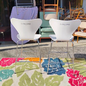 K&ouml;nnte beinhalten: Zwei wei&szlig;e Barhocker mit dem "Lanc&ocirc;me"-Logo auf der R&uuml;ckenlehne. Die Hocker haben ein modernes Design mit verchromten Beinen. Sie stehen auf einem bunten Blumengemusterten Teppich mit rosa, blauen und gr&uuml;nen Blumen.