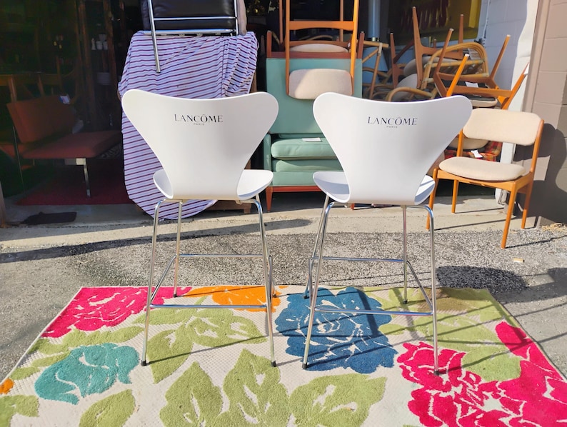 K&ouml;nnte beinhalten: Zwei wei&szlig;e Barhocker mit dem "Lanc&ocirc;me"-Logo auf der R&uuml;ckenlehne. Die Hocker haben ein modernes Design mit verchromten Beinen. Sie stehen auf einem bunten Blumengemusterten Teppich mit rosa, blauen und gr&uuml;nen Blumen.
