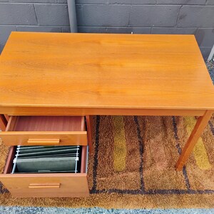 Op de afbeelding: Een rechthoekig houten bureau met een gladde, lichtbruine afwerking en zichtbare houtnerf. Het bureau heeft twee gestapelde lades aan de linkerkant, waarbij de onderste lade hangmappen bevat. Het bureau staat op vier rechte poten.