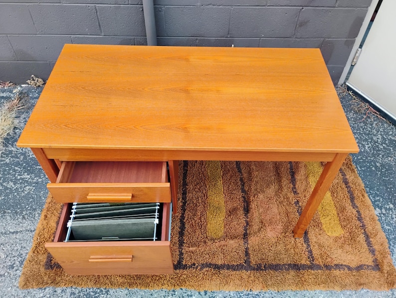 Op de afbeelding: Een rechthoekig houten bureau met een gladde, lichtbruine afwerking en zichtbare houtnerf. Het bureau heeft twee gestapelde lades aan de linkerkant, waarbij de onderste lade hangmappen bevat. Het bureau staat op vier rechte poten.
