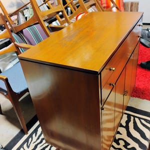 Puede incluir: Un armario de madera vintage con dos puertas y dos cajones. El armario tiene un acabado en madera clara y herrajes de lat&oacute;n. El armario est&aacute; sobre una alfombra de cebra de rayas blancas y negras.