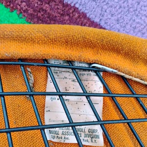 May include: An orange textured fabric seat cushion attached to a black metal wire grid frame. A white rectangular tag is secured to the frame, featuring printed text that reads Knoll Associates Division, 320 Park Ave., New York, N.Y.