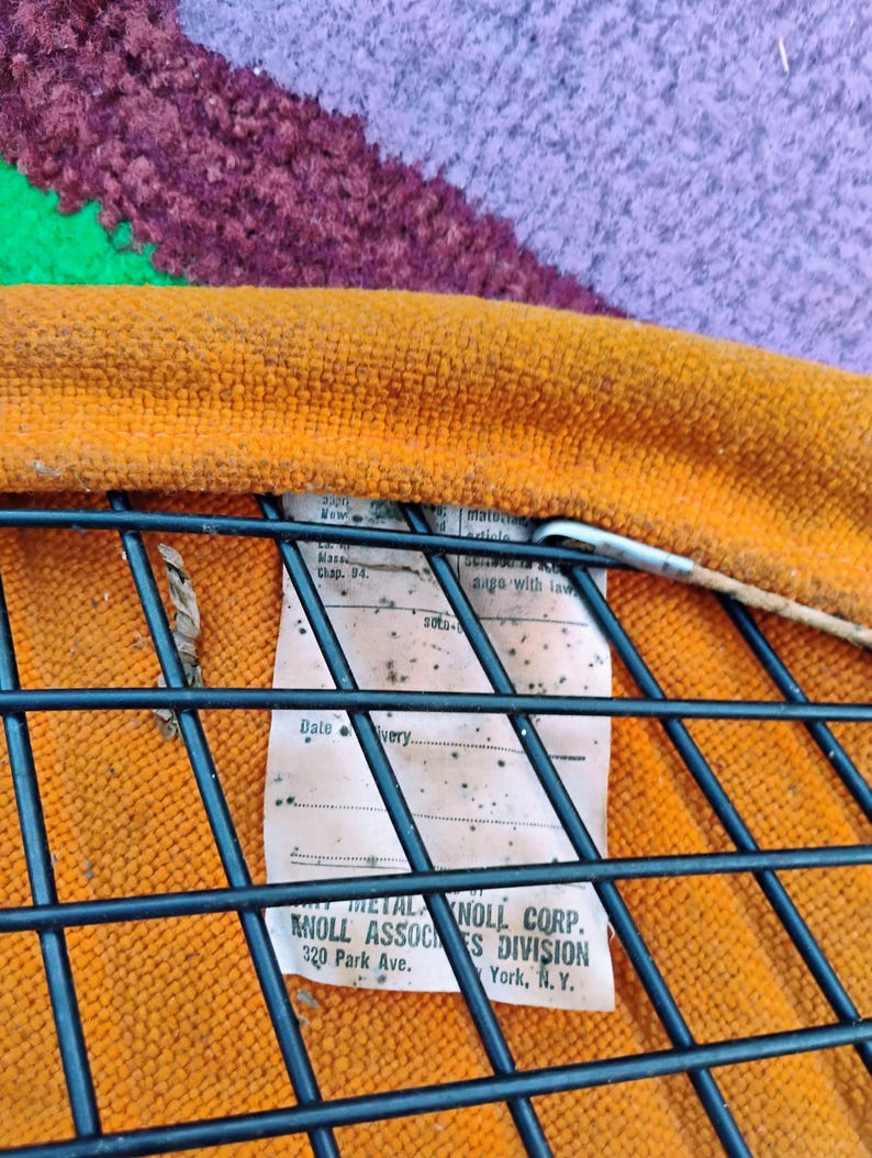 May include: An orange textured fabric seat cushion attached to a black metal wire grid frame. A white rectangular tag is secured to the frame, featuring printed text that reads Knoll Associates Division, 320 Park Ave., New York, N.Y.