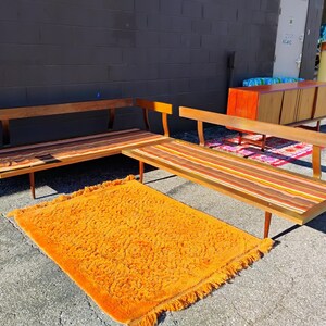 May include: Mid-century modern furniture set featuring a wooden sofa and a matching bench with colorful striped cushions. An orange shag rug is in front of the furniture. The sofa and bench have wooden legs.