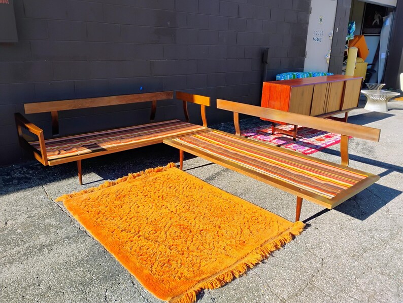 May include: Mid-century modern furniture set featuring a wooden sofa and a matching bench with colorful striped cushions. An orange shag rug is in front of the furniture. The sofa and bench have wooden legs.