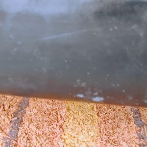 May include: A dark, smooth, and reflective surface with a subtle gradient of gray and brown tones, positioned above a textured, shaggy rug featuring alternating vertical stripes of light orange, tan, and dark brown fibers.