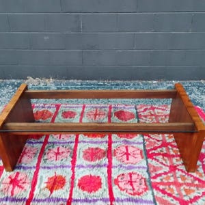 May include: A mid-century modern coffee table with a rectangular wooden frame and a glass top. The table is made of light brown wood and sits on a colorful patterned rug. The table measures approximately 48 inches long.