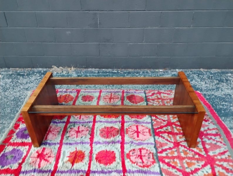 May include: A mid-century modern coffee table with a rectangular wooden frame and a glass top. The table is made of light brown wood and sits on a colorful patterned rug. The table measures approximately 48 inches long.