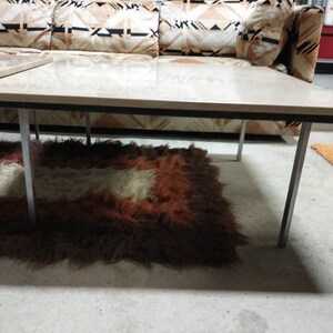May include: A mid-century modern coffee table with a light wood top and chrome legs. The table is rectangular and sits on a shaggy rug with brown, white, and orange tones. A patterned sofa is in the background.