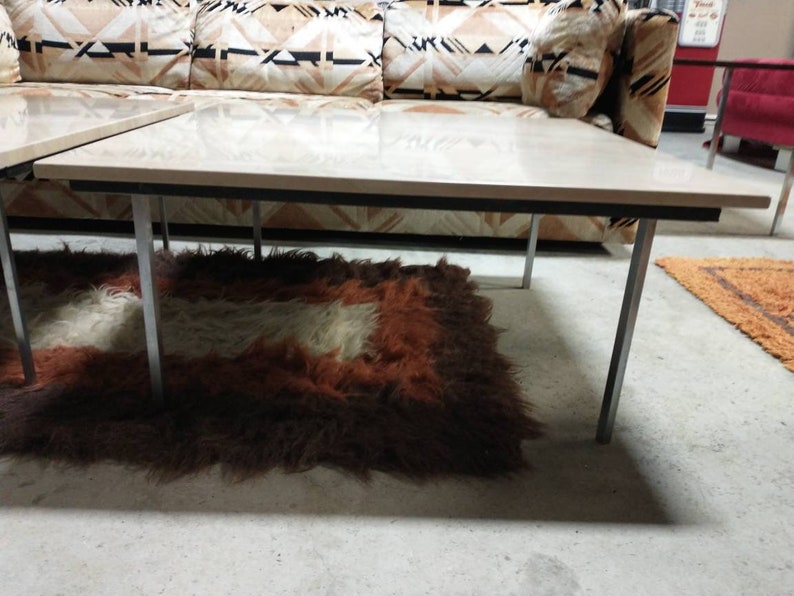 May include: A mid-century modern coffee table with a light wood top and chrome legs. The table is rectangular and sits on a shaggy rug with brown, white, and orange tones. A patterned sofa is in the background.
