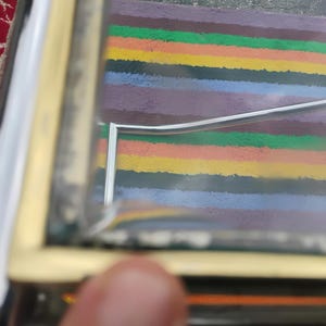May include: A rectangular glass display box featuring a metallic gold-toned frame. The structure includes clear glass panels and a hinged lid. The interior base is lined with a vibrant, multi-colored striped pattern in shades of orange, yellow, green, and blue.