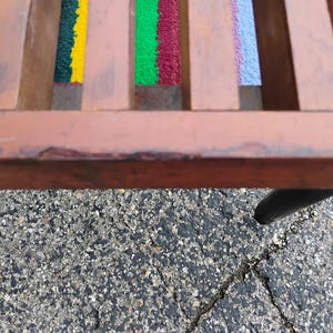 K&ouml;nnte beinhalten: Nahaufnahme einer Holzbank mit farbenfrohen Webakzenten. Die Bank hat einen braunen Rahmen und schwarze Beine. Die gewebten Abschnitte weisen gelbe, gr&uuml;ne, rote und blaue Farbt&ouml;ne auf und verleihen dem Design eine lebendige Note.