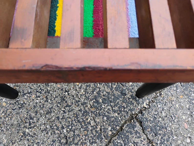 K&ouml;nnte beinhalten: Nahaufnahme einer Holzbank mit farbenfrohen Webakzenten. Die Bank hat einen braunen Rahmen und schwarze Beine. Die gewebten Abschnitte weisen gelbe, gr&uuml;ne, rote und blaue Farbt&ouml;ne auf und verleihen dem Design eine lebendige Note.