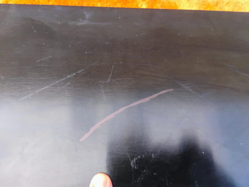 May include: A flat, black, rectangular surface with a smooth, reflective finish. The surface displays several visible light-colored scratches and a prominent, thin, diagonal mark across the center, indicating wear on the material's dark coating.