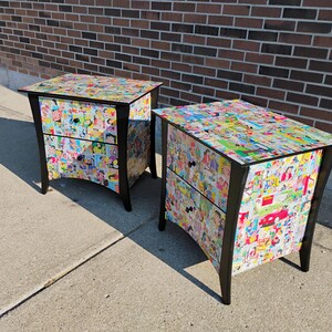 May include: Two black nightstands with two drawers each. The nightstands are covered in a collage of colorful comic book images. The nightstands have black legs and black drawer pulls. The nightstands are on a concrete surface.