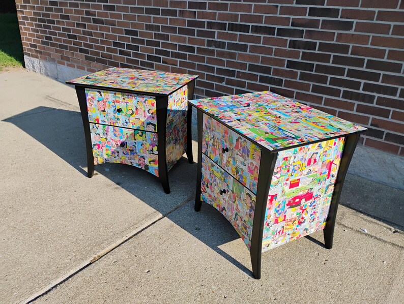 May include: Two black nightstands with two drawers each. The nightstands are covered in a collage of colorful comic book images. The nightstands have black legs and black drawer pulls. The nightstands are on a concrete surface.