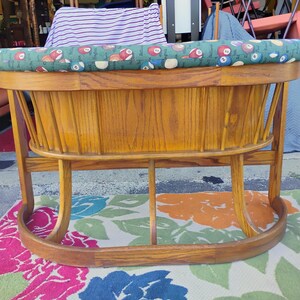 May include: A vintage wooden rocking cradle with a green and patterned cushion. The cradle has a curved base and a slatted wooden body. The cushion features a pattern of billiard balls. The cradle is made of light brown wood.