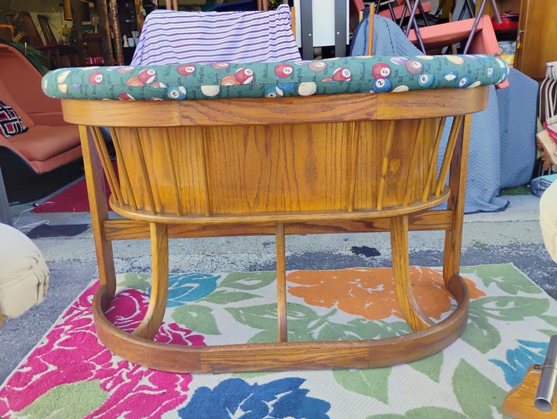 May include: A vintage wooden rocking cradle with a green and patterned cushion. The cradle has a curved base and a slatted wooden body. The cushion features a pattern of billiard balls. The cradle is made of light brown wood.