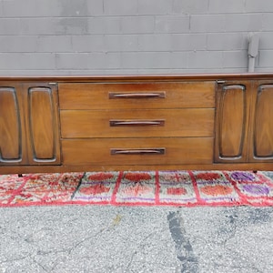 May include: A long, rectangular wooden sideboard featuring a warm brown finish. The piece includes three central horizontal drawers with dark, elongated handles, flanked by two cabinet doors on either side, each with a recessed vertical panel design.