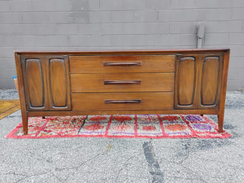 May include: A long, rectangular wooden sideboard featuring a warm brown finish. The piece includes three central horizontal drawers with dark, elongated handles, flanked by two cabinet doors on either side, each with a recessed vertical panel design.