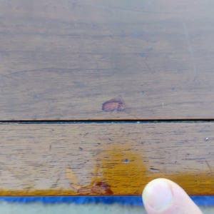 May include: Close-up of a wooden surface with a small red stain. The surface is brown and has a natural wood grain. The surface is part of a piece of furniture.