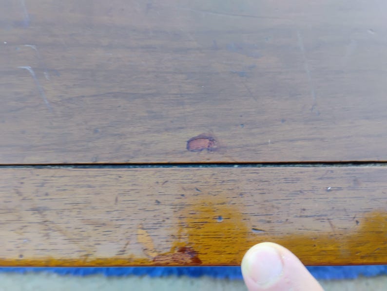 May include: Close-up of a wooden surface with a small red stain. The surface is brown and has a natural wood grain. The surface is part of a piece of furniture.