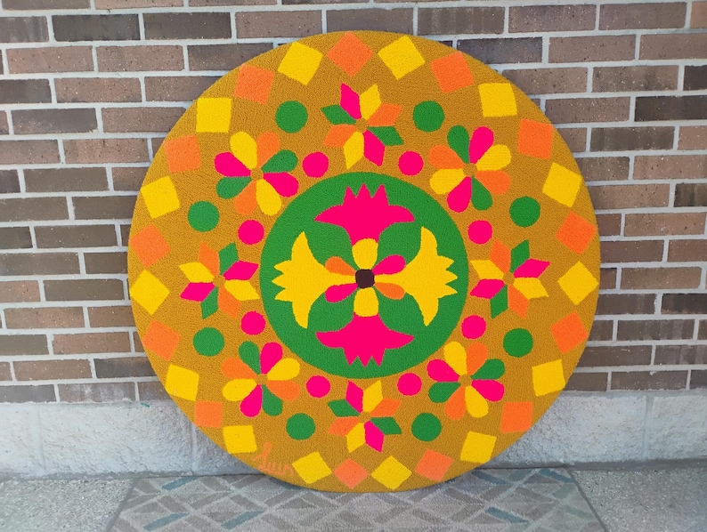 Puede incluir: Una alfombra grande, redonda y colorida con un dise&ntilde;o floral geom&eacute;trico. La alfombra presenta un fondo amarillo mostaza con formas florales y geom&eacute;tricas naranjas, amarillas, verdes y rosas. La alfombra est&aacute; colocada sobre una superficie estampada.