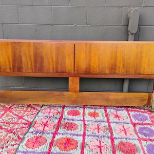 May include: A rectangular wooden headboard featuring a warm brown wood grain finish. The headboard consists of a solid upper panel divided by a vertical seam and a lower horizontal support bar, with metallic mounting brackets attached at both ends.