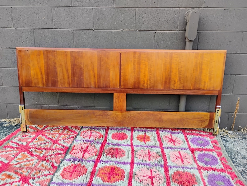 May include: A rectangular wooden headboard featuring a warm brown wood grain finish. The headboard consists of a solid upper panel divided by a vertical seam and a lower horizontal support bar, with metallic mounting brackets attached at both ends.