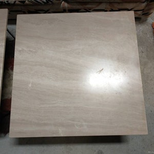 May include: Two square, light beige stone tables with a marbled appearance. The tables have a smooth surface and are likely made of travertine or a similar material. The tables are side tables or coffee tables.