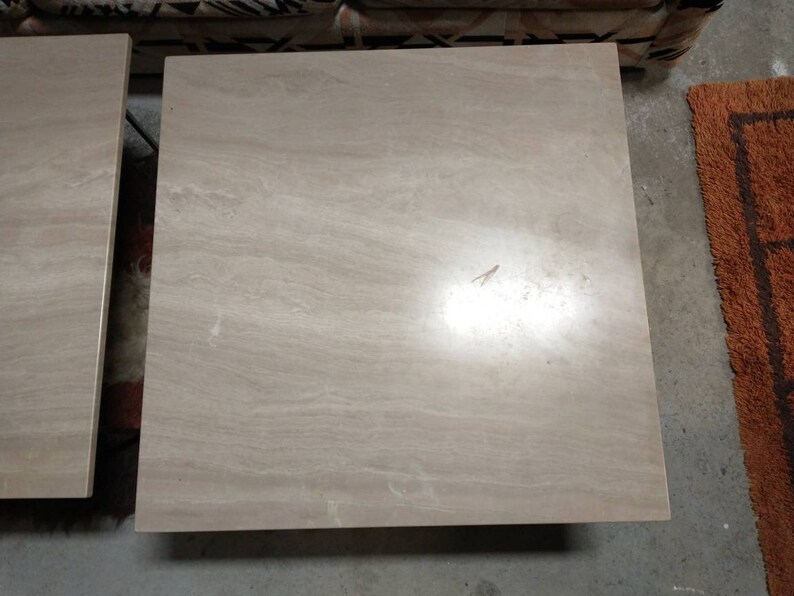 May include: Two square, light beige stone tables with a marbled appearance. The tables have a smooth surface and are likely made of travertine or a similar material. The tables are side tables or coffee tables.