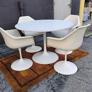 May include: A white circular dining table with a central pedestal base, accompanied by four matching white armchairs, each featuring a curved shell seat and a single pedestal base, arranged in a set for dining or seating purposes.