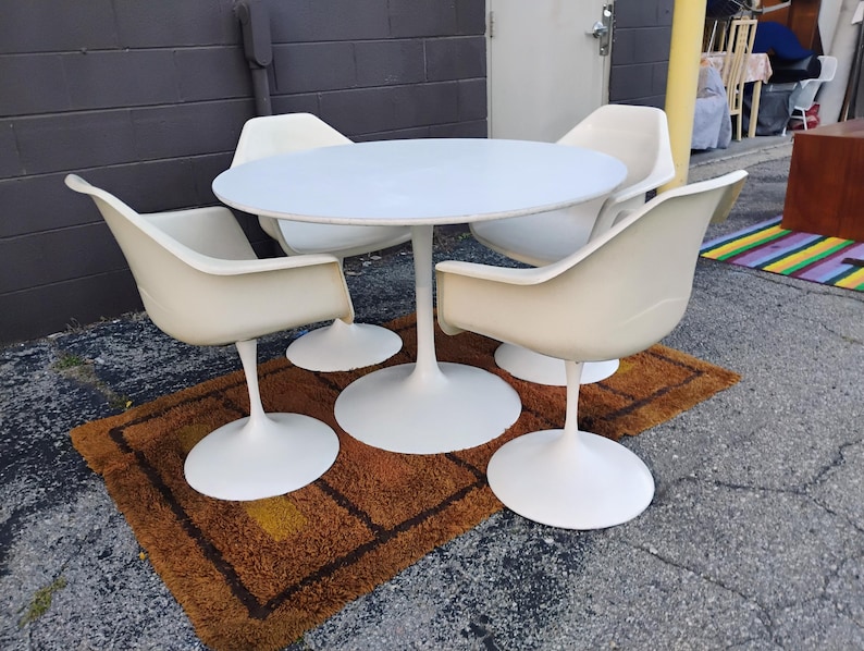 May include: A white circular dining table with a central pedestal base, accompanied by four matching white armchairs, each featuring a curved shell seat and a single pedestal base, arranged in a set for dining or seating purposes.