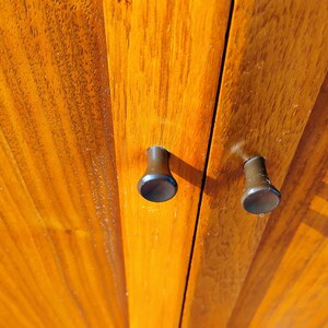 Op de afbeelding: Twee kleine, donkerbruine, cilindervormige kastknoppen met platte ronde uiteinden zijn gemonteerd op een verticaal houten paneel. Het hout heeft een warme, goudbruine tint met een zichtbare, rechte houtnerf die verticaal loopt.