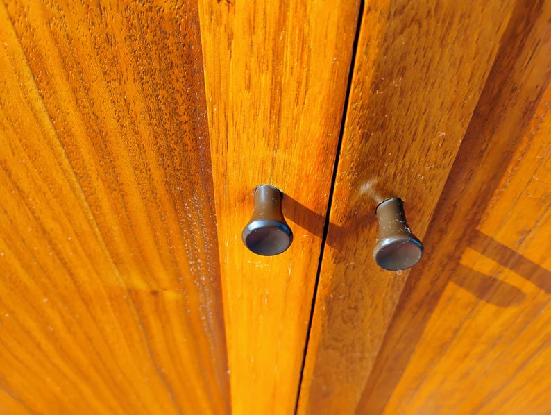 Op de afbeelding: Twee kleine, donkerbruine, cilindervormige kastknoppen met platte ronde uiteinden zijn gemonteerd op een verticaal houten paneel. Het hout heeft een warme, goudbruine tint met een zichtbare, rechte houtnerf die verticaal loopt.