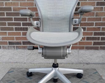 Könnte beinhalten: Ein hellgrauer ergonomischer Bürostuhl mit atmungsaktiver Netzrückenlehne und Sitzfläche, verstellbaren Armlehnen und einem Fünf-Punkt-Fußkreuz mit Rollen. Der Stuhl verfügt über einen stabilen Rahmen mit mechanischen Einstellhebeln.