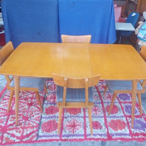 May include: A set of four mid-century modern dining chairs with a light wood frame and gray fabric seats. The chairs are arranged around a rectangular table with a light wood top. The table and chairs are on a red and purple rug.
